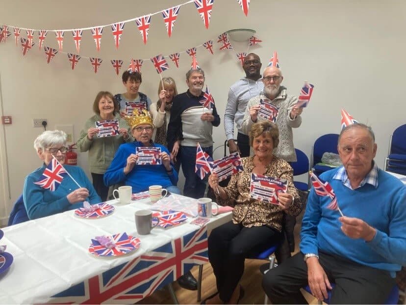 A group of people celebrating with British flags and decorations, seated and standing around a table, smiling for the camera. - Home Instead