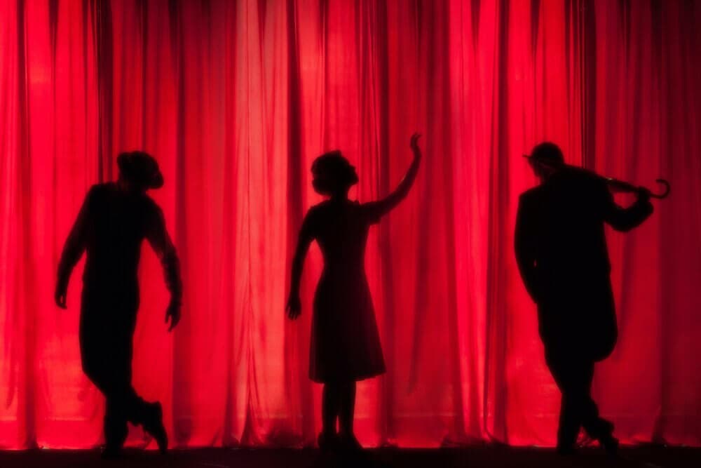 Three silhouetted figures pose dramatically against a red curtain, one holding an umbrella aloft. - Home Instead