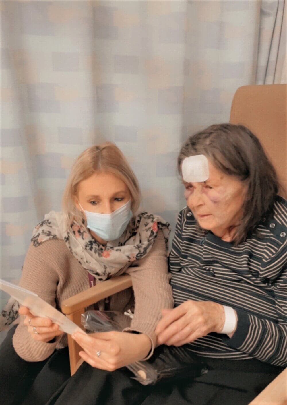 A woman wearing a mask sits beside an elderly woman with bandages, looking at papers together in a room. - Home Instead