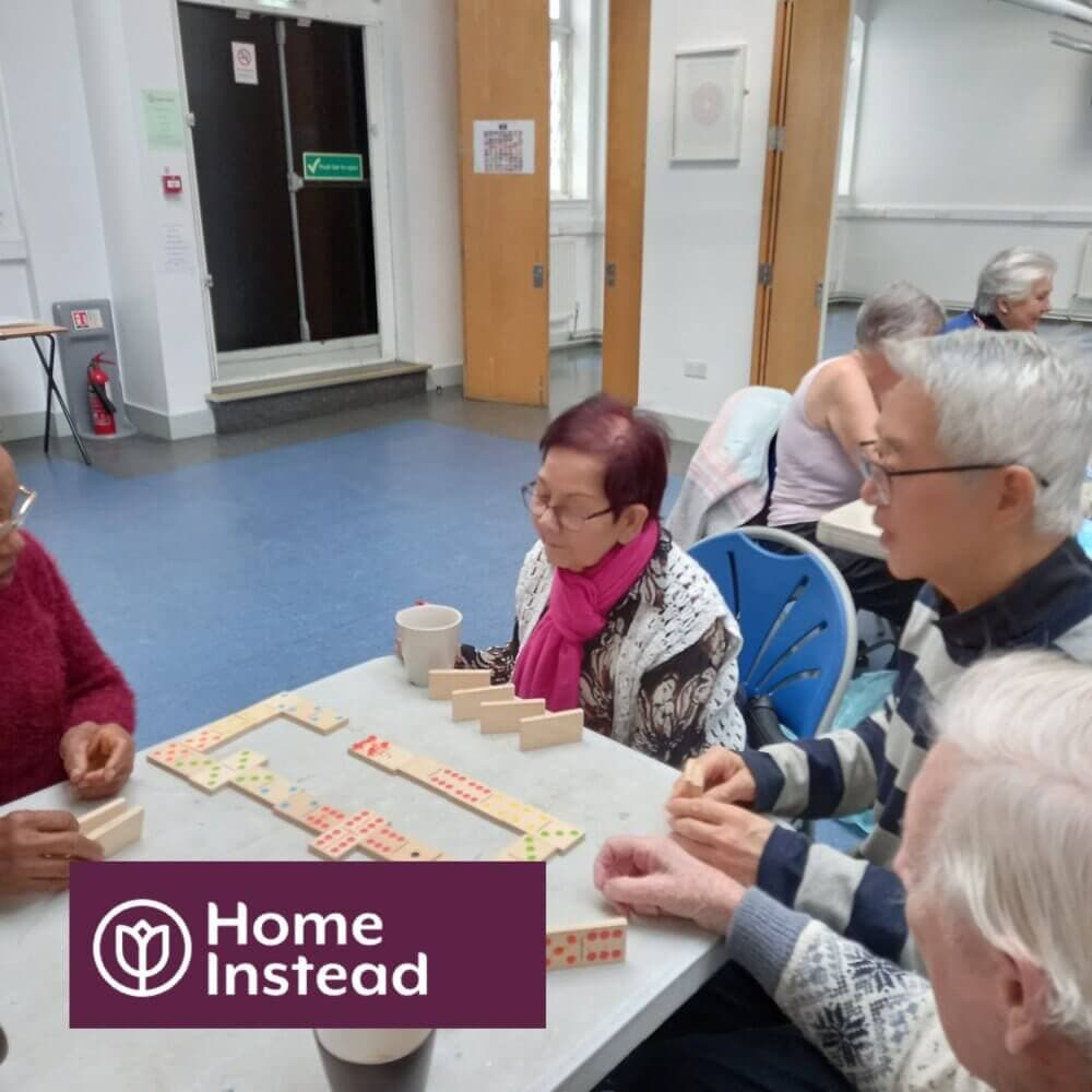 Seniors gathered around a table playing dominoes in a community center, with a "Home Instead" logo at the bottom left. - Home Instead