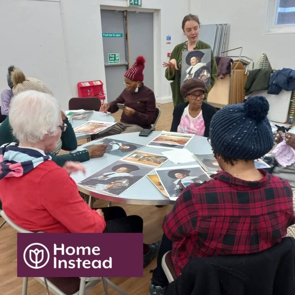 A group of elderly people are seated around a table engaging in an art activity with a caregiver standing nearby. - Home Instead