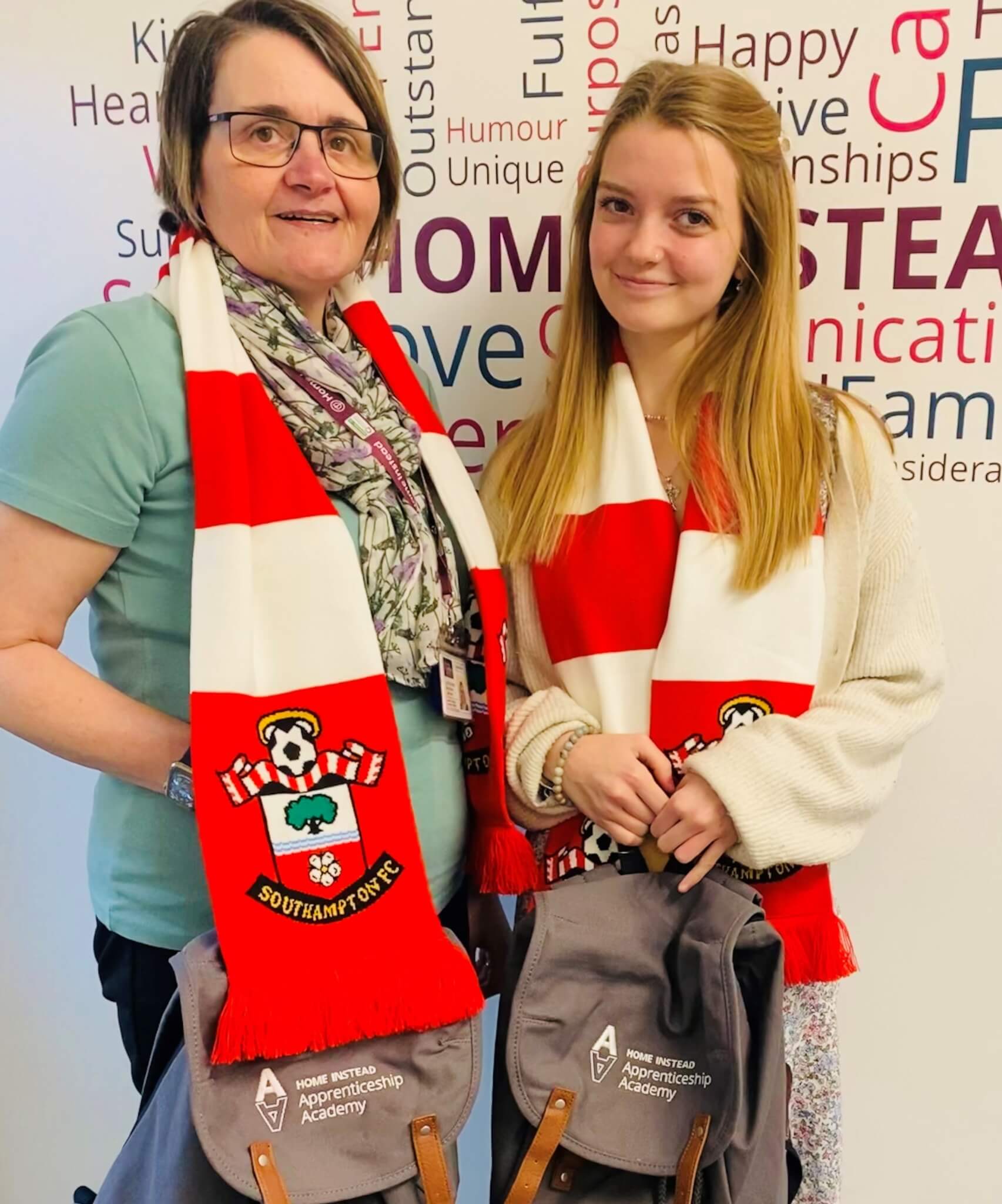 Two women wearing Southampton F.C. scarves, holding grey bags, posing in front of a wall with motivational words. - Home Instead