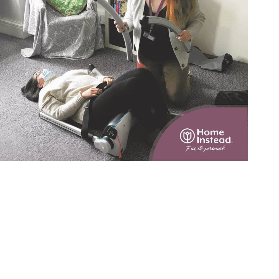 A caregiver assists a person lying on a mobility device at home with a "Home Instead" logo visible in the corner. - Home Instead