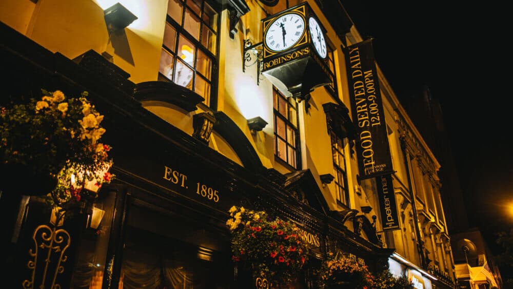A warmly lit, historic pub building with a hanging clock and flowers, established in 1895, advertising daily food service. - Home Instead