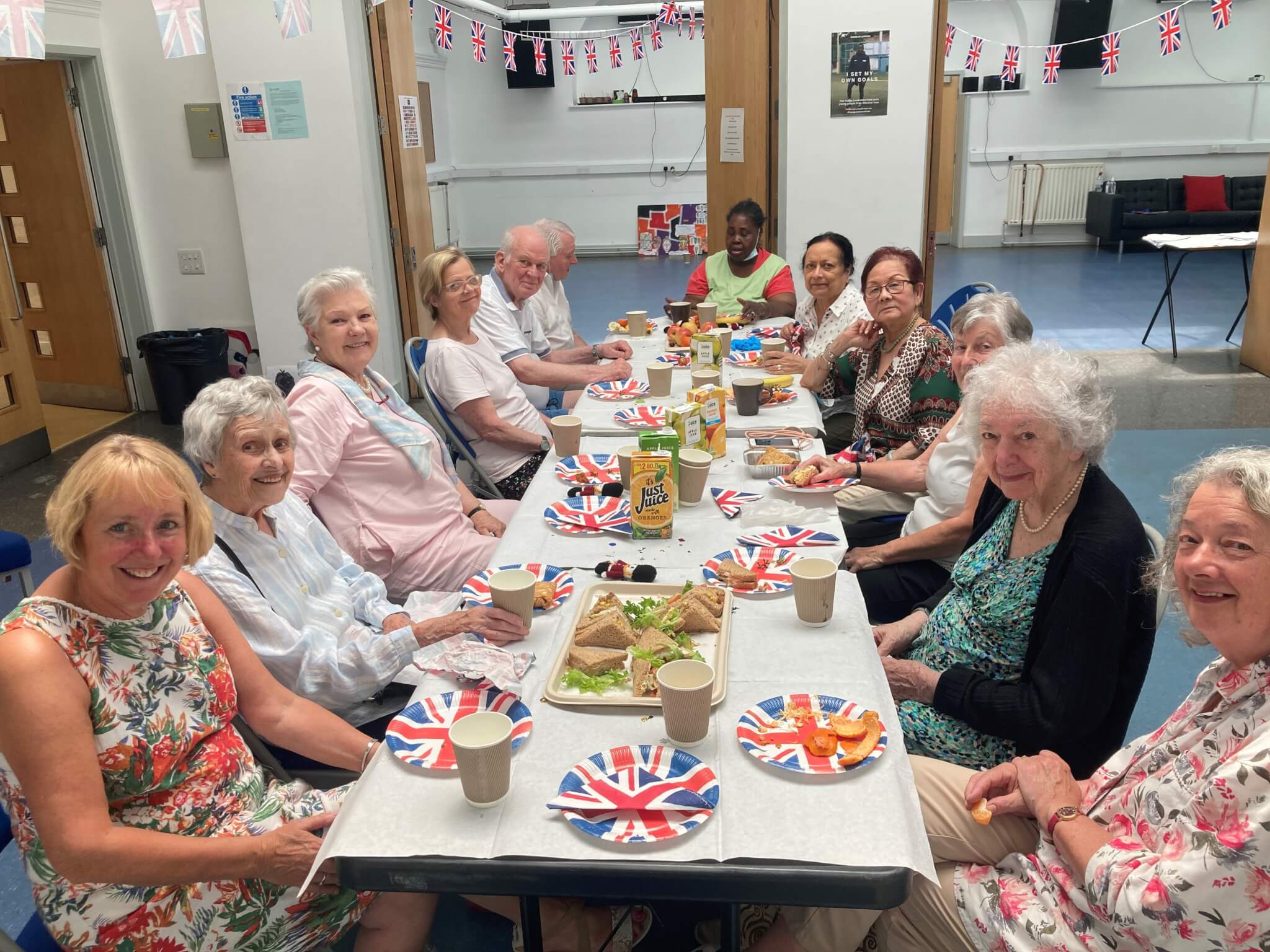 A group of elderly people smile around a table set with food and Union Jack-themed decorations. - Home Instead