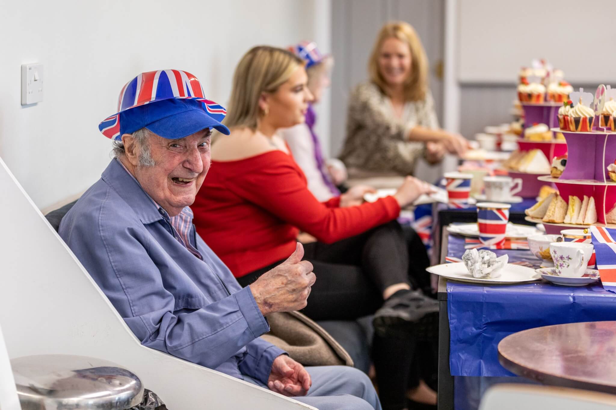 Elderly man in a Union Jack hat and jacket gives a thumbs up at a festive tea party with others in the background. - Home Instead