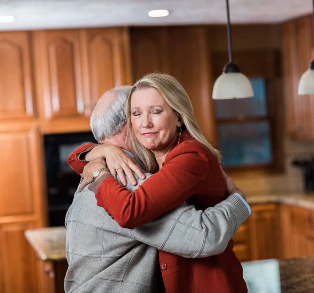 Two people embrace warmly in a kitchen with wooden cabinets in the background. - Home Instead