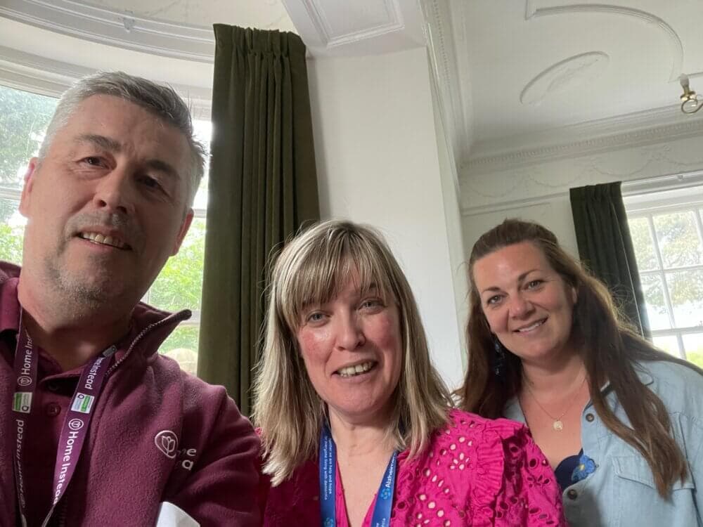 Three people smiling indoors, with large windows and green curtains in the background. - Home Instead
