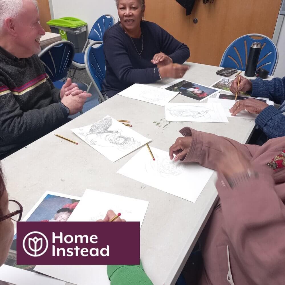 A group of people engaged in art activities around a table with "Home Instead" logo in the corner. - Home Instead