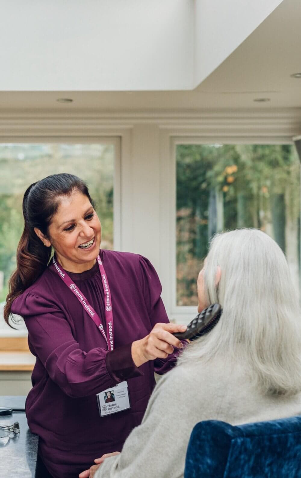 A caregiver wearing a purple shirt is brushing the hair of an older woman with long white hair in a bright room. - Home Instead Bournemouth & Christchurch