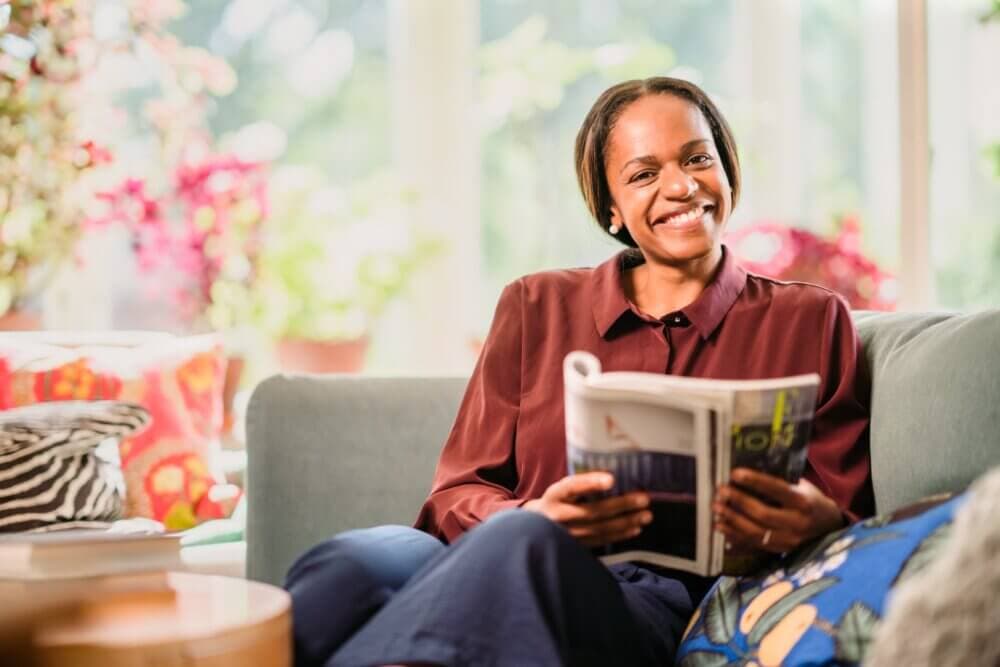 Smiling woman sitting on a couch reading a magazine in a brightly lit, cosy living room with colourful decor. - Home Instead Poole