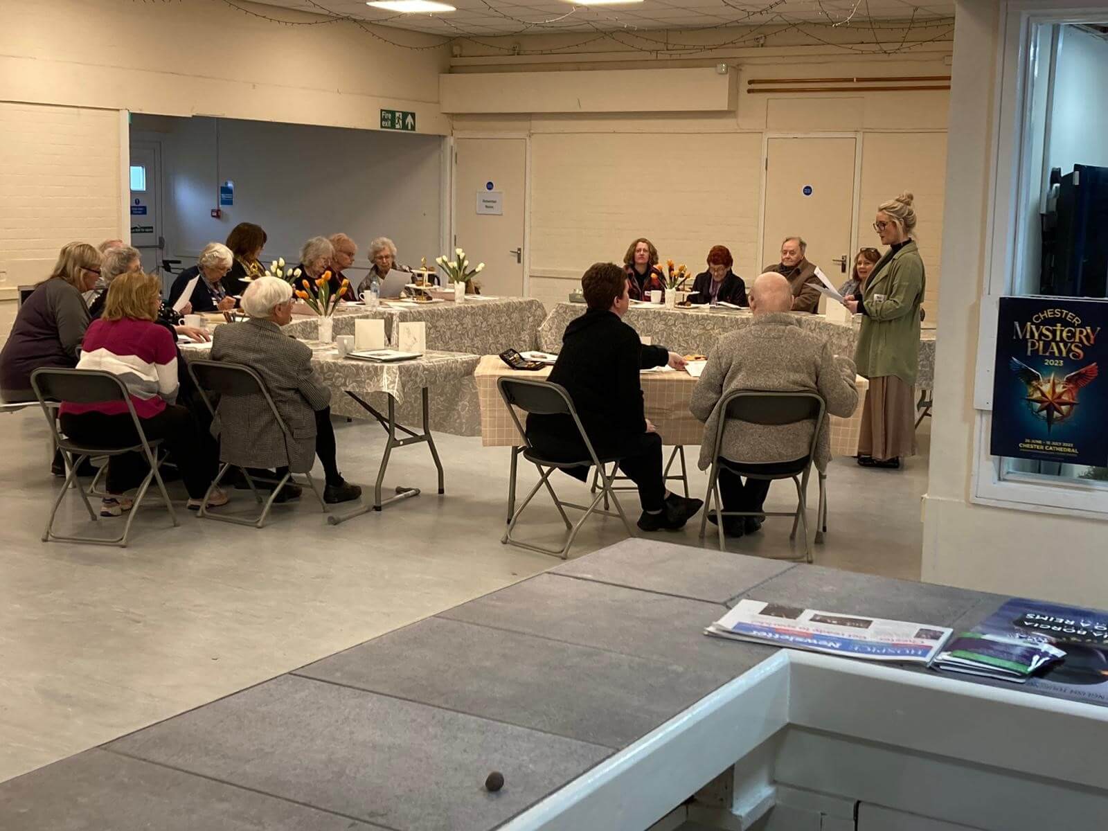People seated around tables in a room, attending a meeting or gathering. A person stands speaking at the front. - Home Instead
