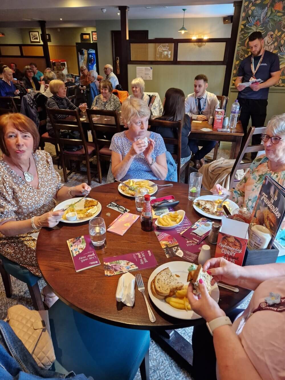 A group of people eating and chatting around a table at a busy restaurant. - Home Instead