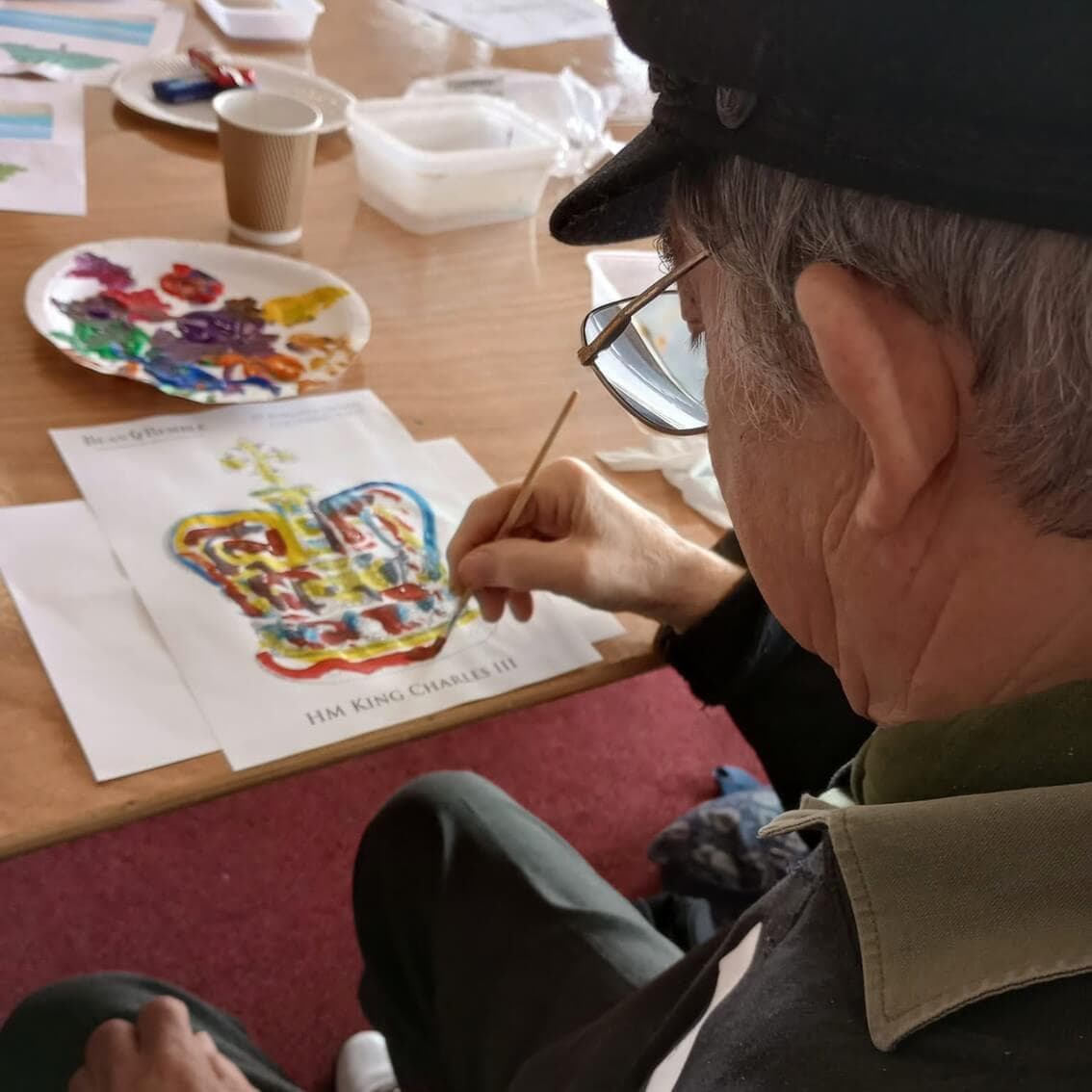 An elderly man wearing a cap and glasses paints a colorful image of a crown at a table. - Home Instead