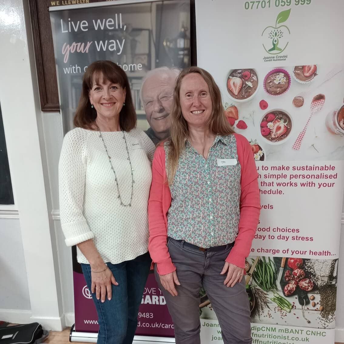 Two women smiling and standing side by side in front of health-related promotional banners. - Home Instead