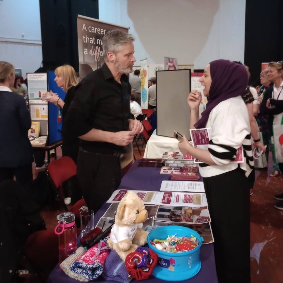 A man and a woman in a headscarf talk at a career fair booth with brochures and toys displayed on the table. - Home Instead