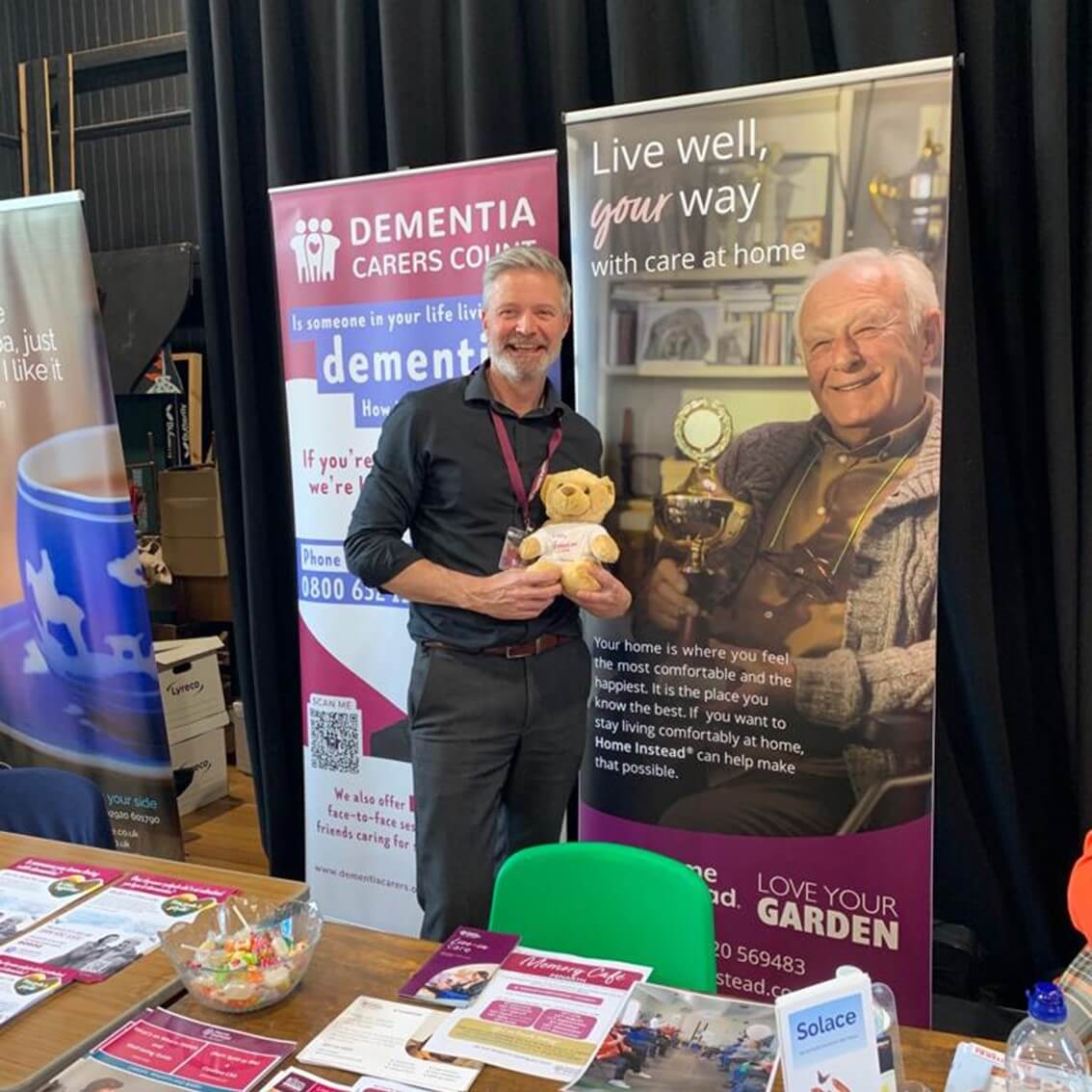 A man holding a teddy bear stands in front of banners about dementia care at an informational booth. - Home Instead