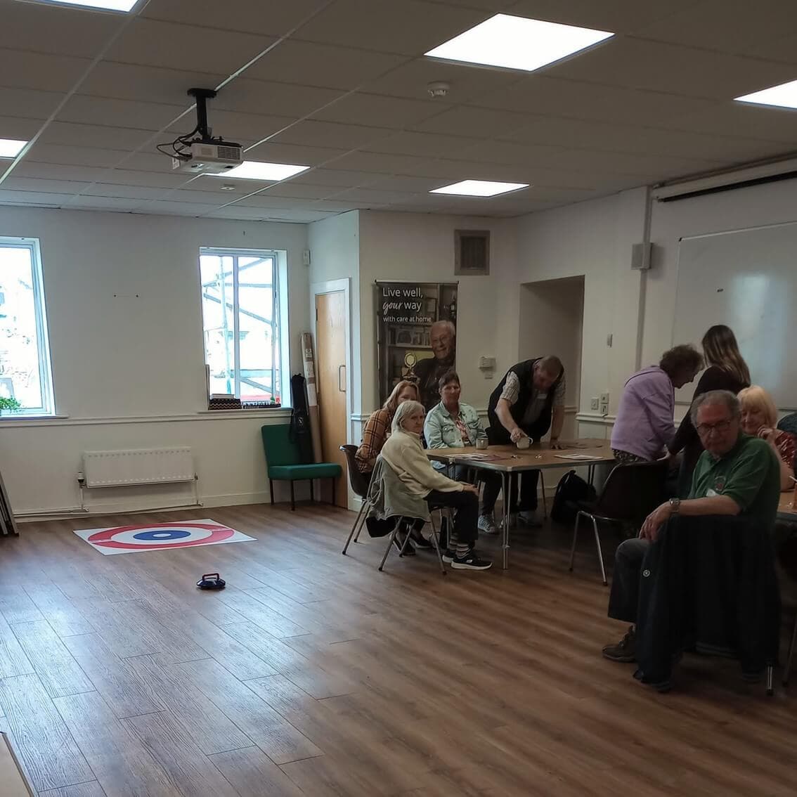 A group of people gather around tables in a room with a curling target mat on the floor. - Home Instead
