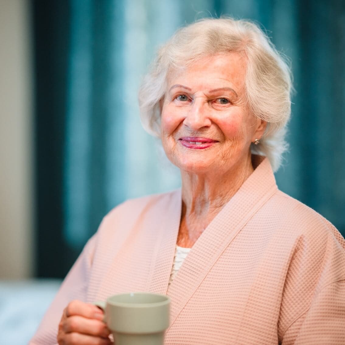 Elderly woman with white hair holding a coffee mug, wearing a light pink robe, smiling warmly. - Home Instead