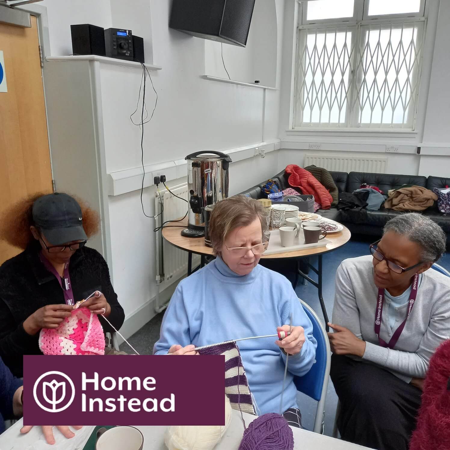 A group of people knitting together in a room, with the Home Instead logo in the bottom left corner. - Home Instead