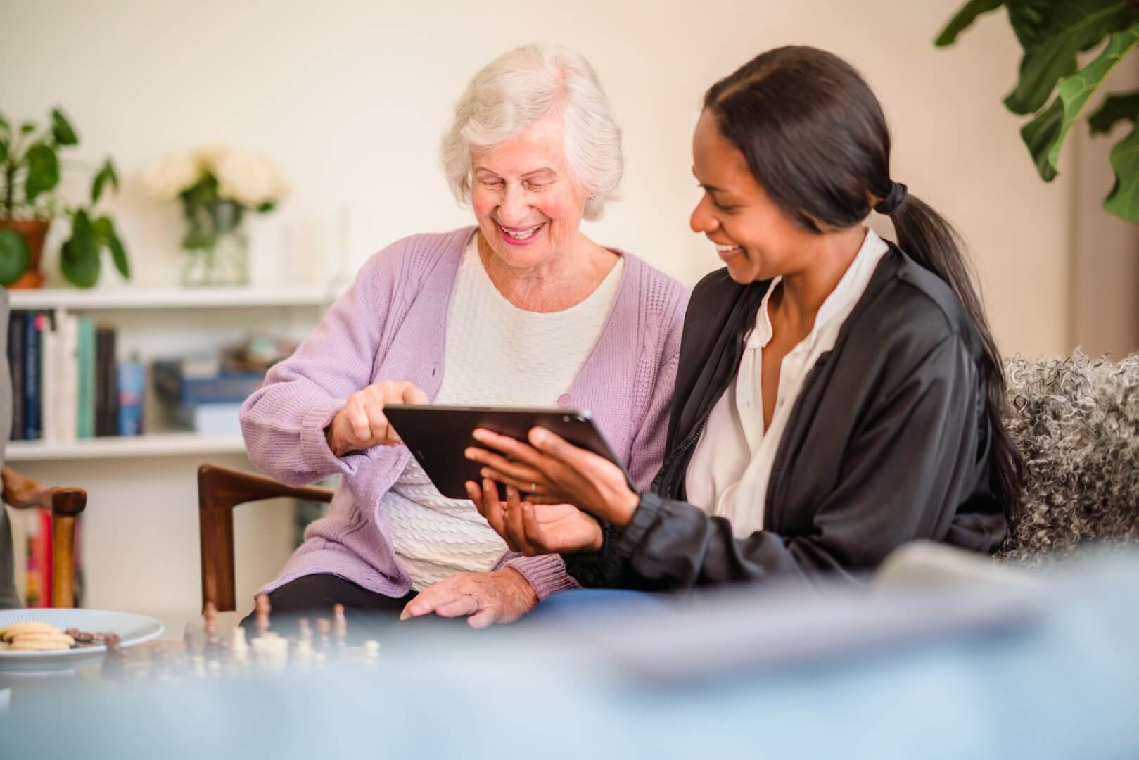 An elderly woman and a younger woman smile while looking at a tablet together in a cozy living room. - Home Instead