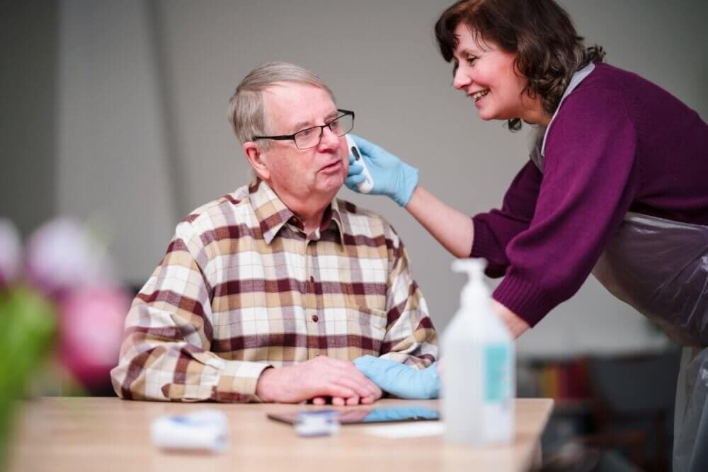 A woman in gloves and apron gently wipes an elderly man's face as he sits at a table in a care setting. - Home Instead