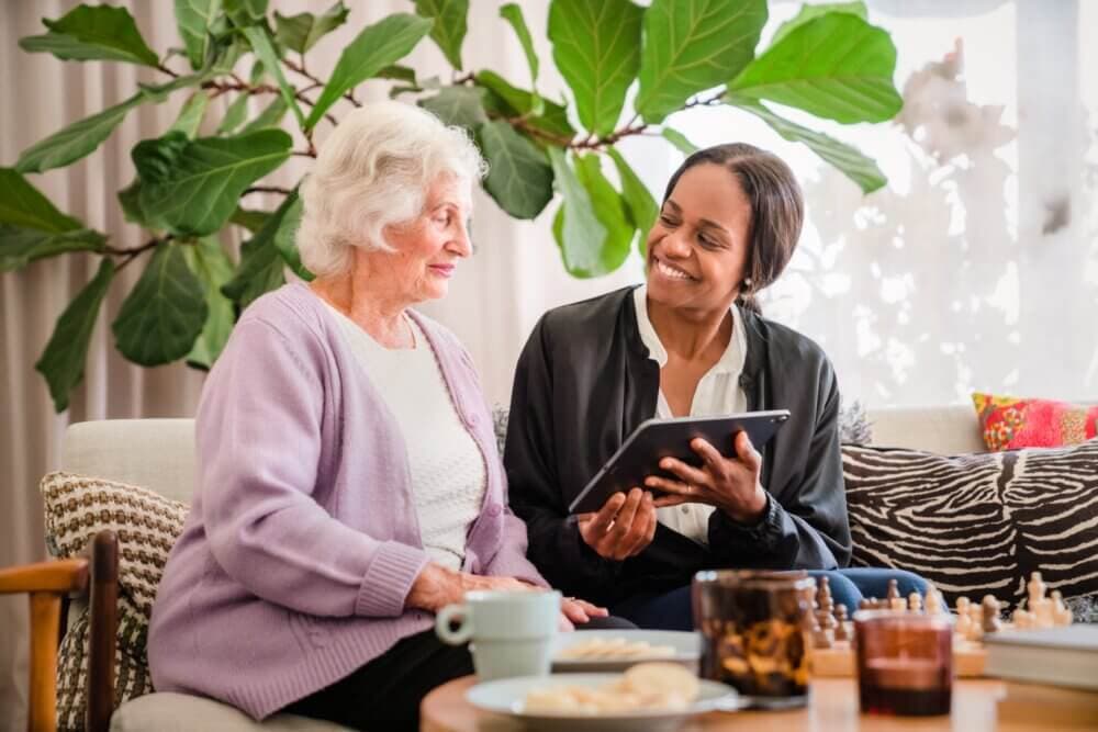 An elderly woman and a younger woman smiling while looking at a tablet together on a cozy sofa with plants in the background. - Home Instead