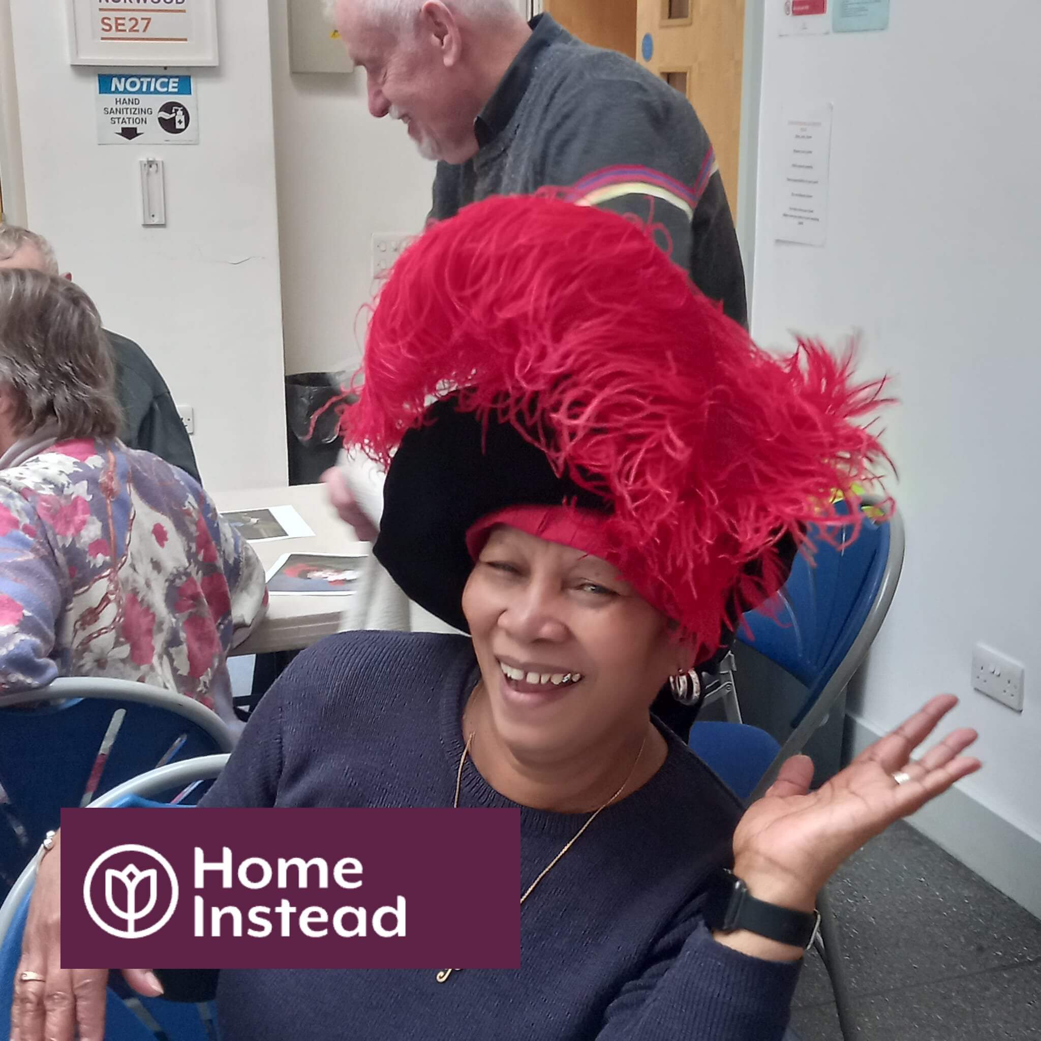 Person wearing a large red feather hat, smiling and waving at a community event. "Home Instead" logo in the corner. - Home Instead
