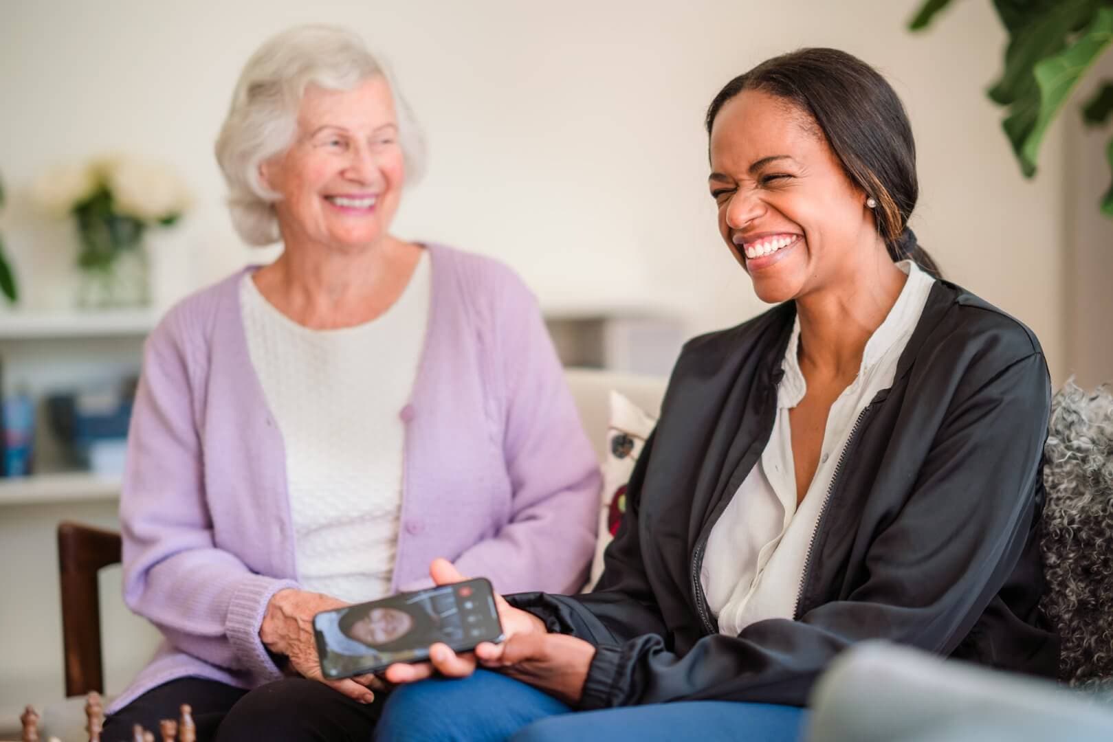 Two women, an elderly woman wearing a purple cardigan and a smiling younger woman holding a phone, sitting together. - Home Instead