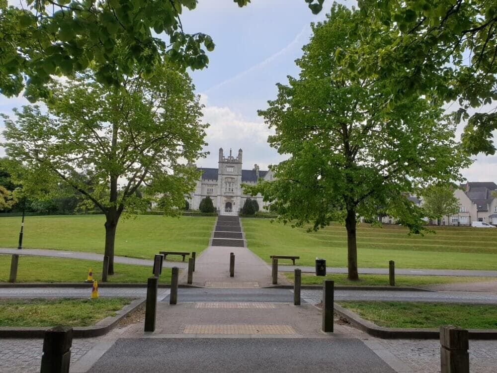 Pathway lined with trees leading to a large historical building with a tower, situated amidst a well-manicured lawn. - Home Instead
