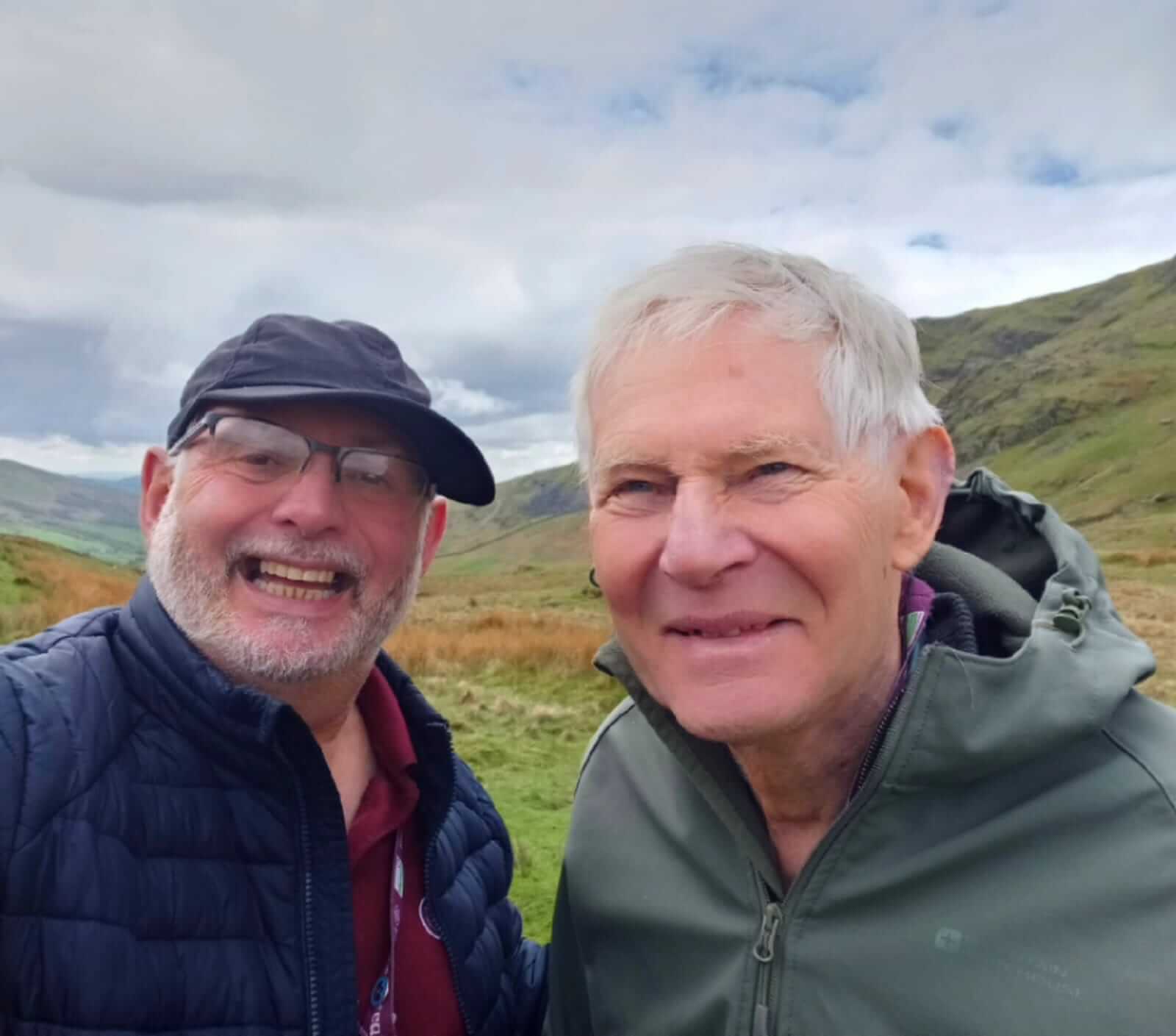 Two elderly men smiling outdoors with hilly landscape and cloudy sky in the background. - Home Instead