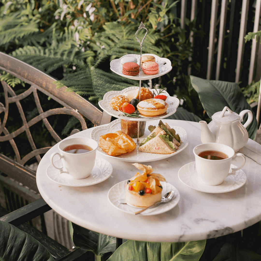 Three-tiered tray with assorted pastries and fruits, two teacups, a teapot, and plates on a marble table in a garden setting. - Home Instead