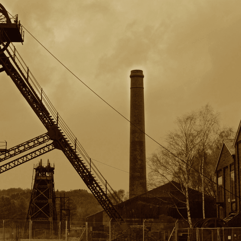 Sepia-toned image of an old industrial site with a tall smokestack, metal structures, and buildings, under a cloudy sky. - Home Instead