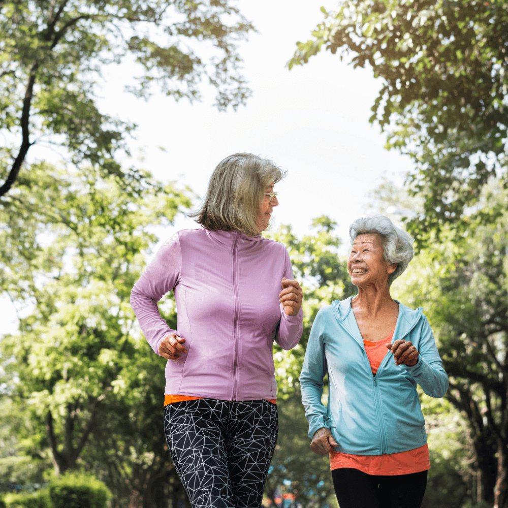 Two elderly women smiling and jogging together in a park surrounded by trees on a sunny day. - Home Instead