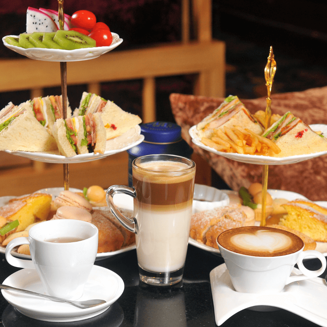 A table with assorted sandwiches, pastries, a latte, a cappuccino, and a cup of tea next to a tiered serving stand. - Home Instead