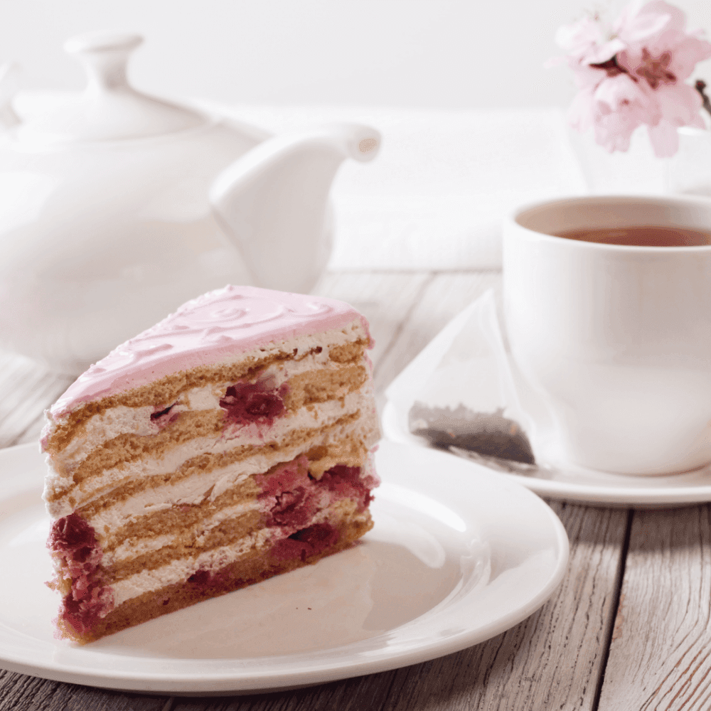 A slice of layered raspberry cake on a plate with a cup of tea and a teapot, set on a white wooden table. - Home Instead
