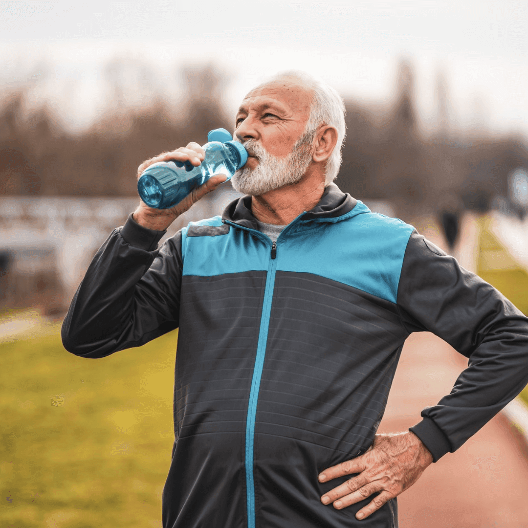 Older man in a tracksuit drinks water from a blue bottle while standing on a running track outdoors. - Home Instead