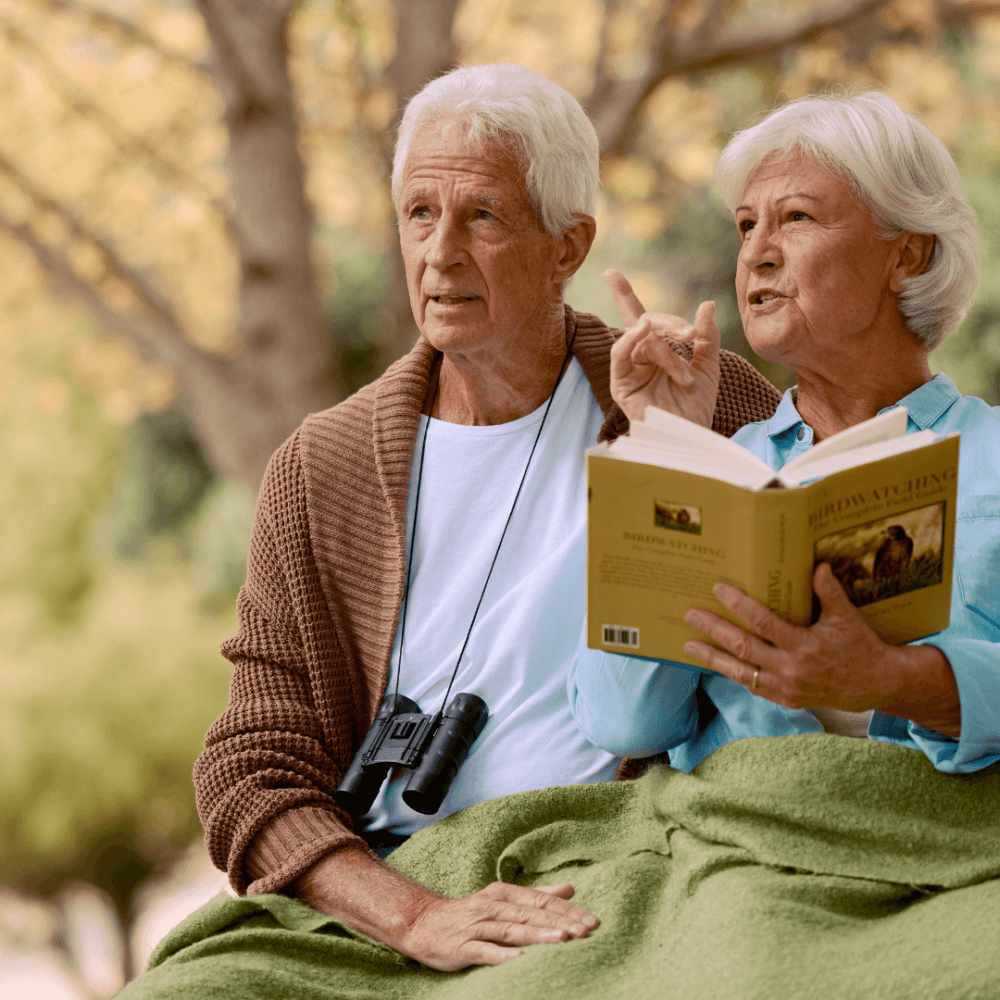 An elderly couple sits on a bench, woman reading a book aloud and man holding binoculars, with trees in the background. - Home Instead
