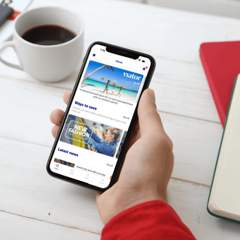 Person holding smartphone displaying a news app; cup of coffee, red notebook, and open book on the white table. - Home Instead