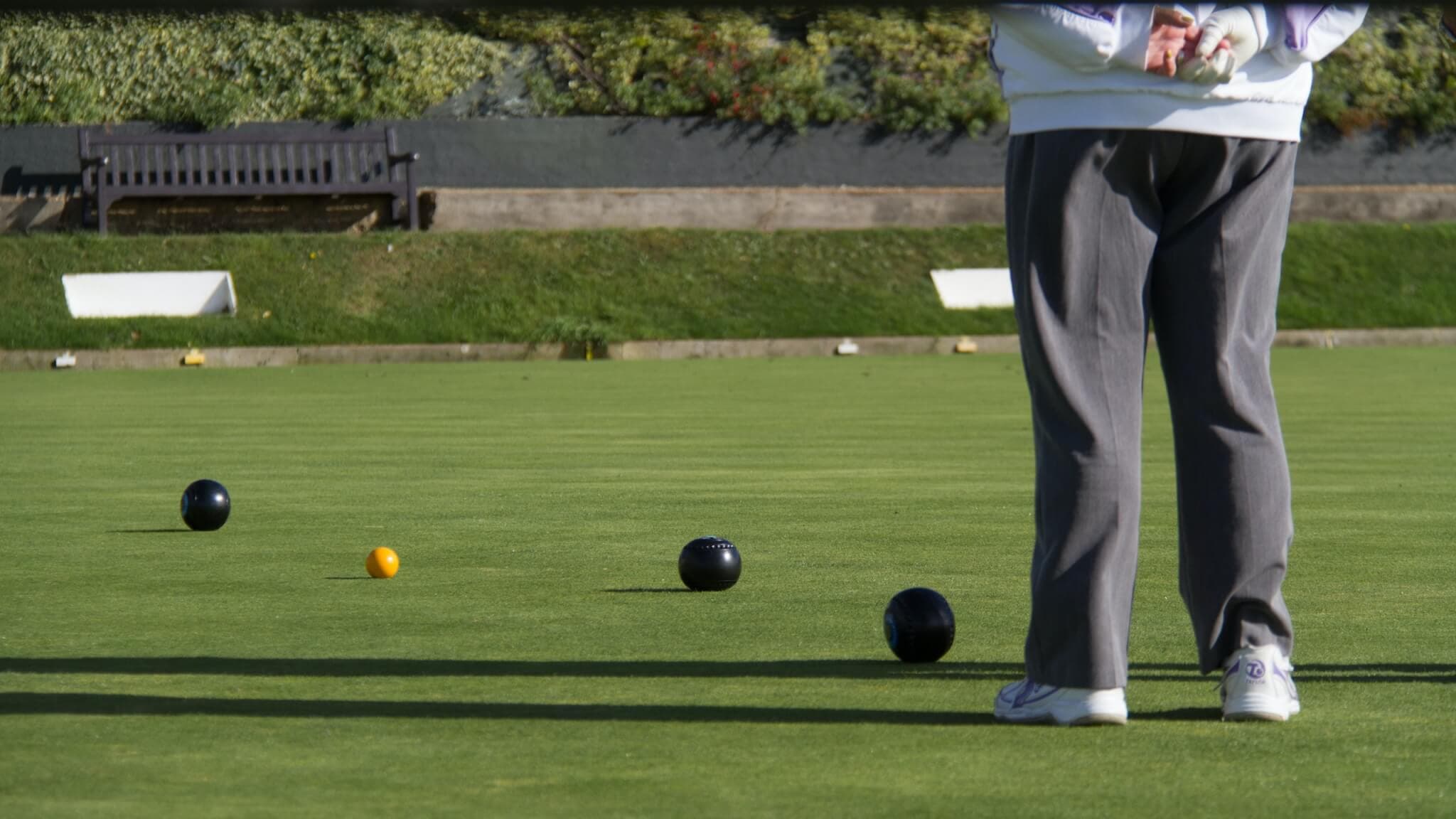 Person in gray pants playing lawn bowls, with several black and yellow bowls on the green field. - Home Instead