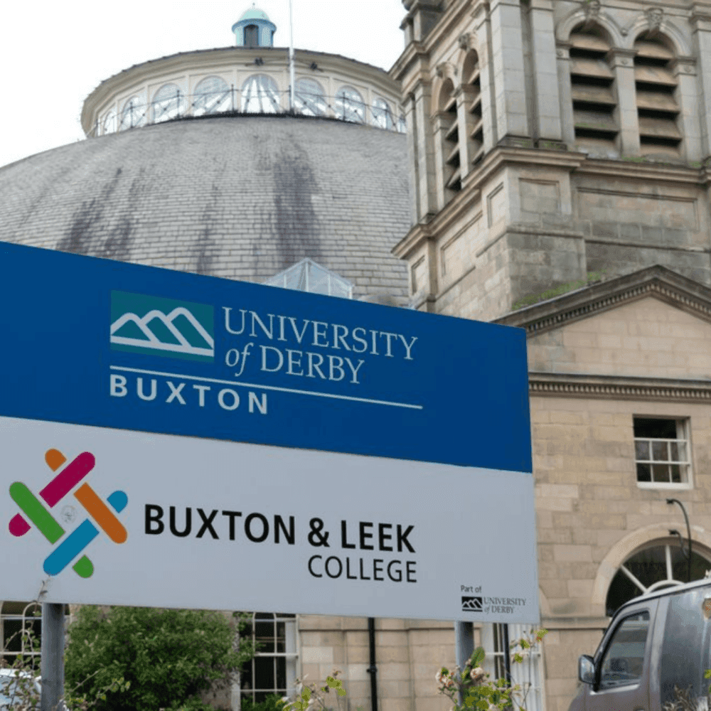 Sign for Buxton & Leek College in front of a University of Derby building with a dome roof in the background. - Home Instead