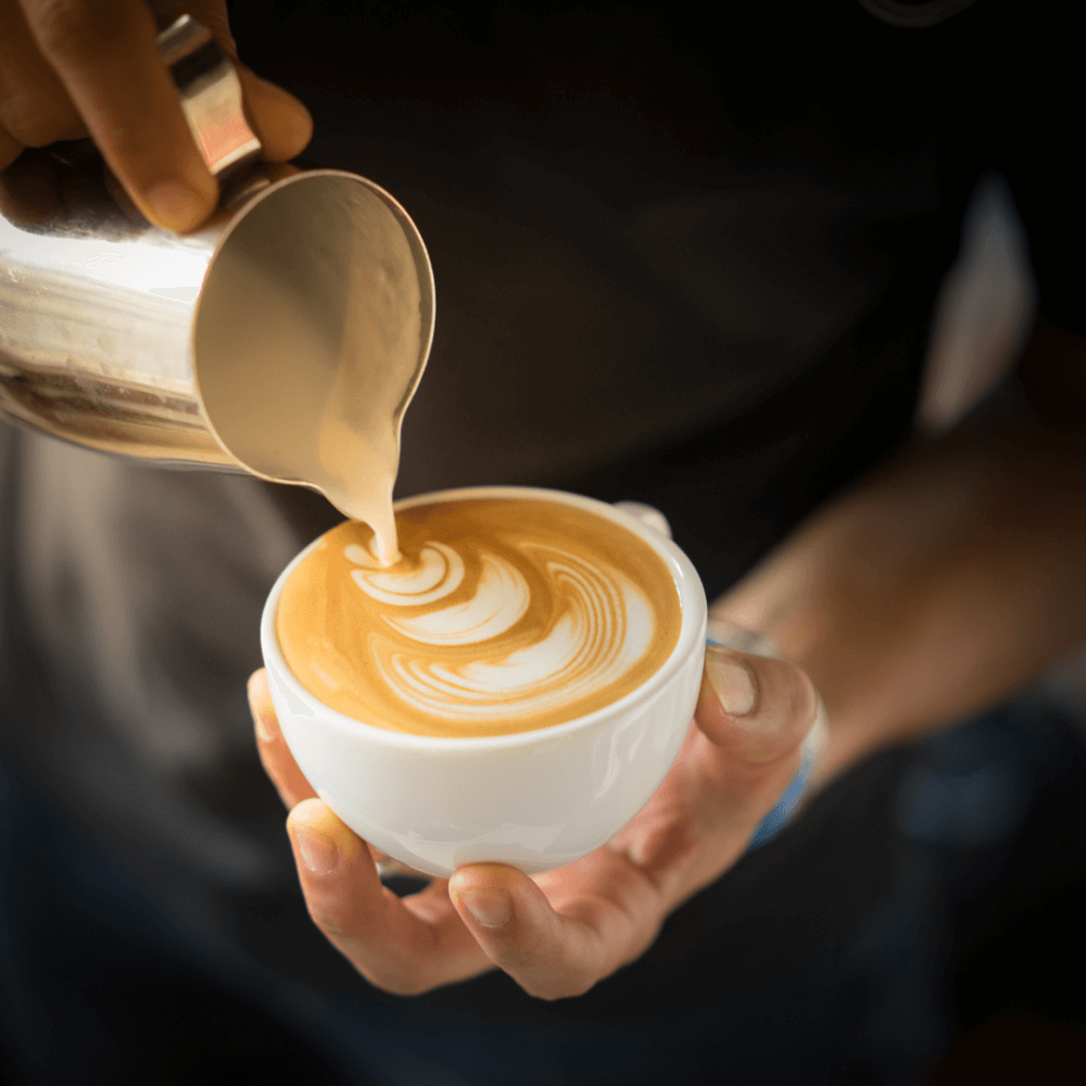 Barista pouring milk into a cup, creating a latte art pattern on a creamy coffee. - Home Instead