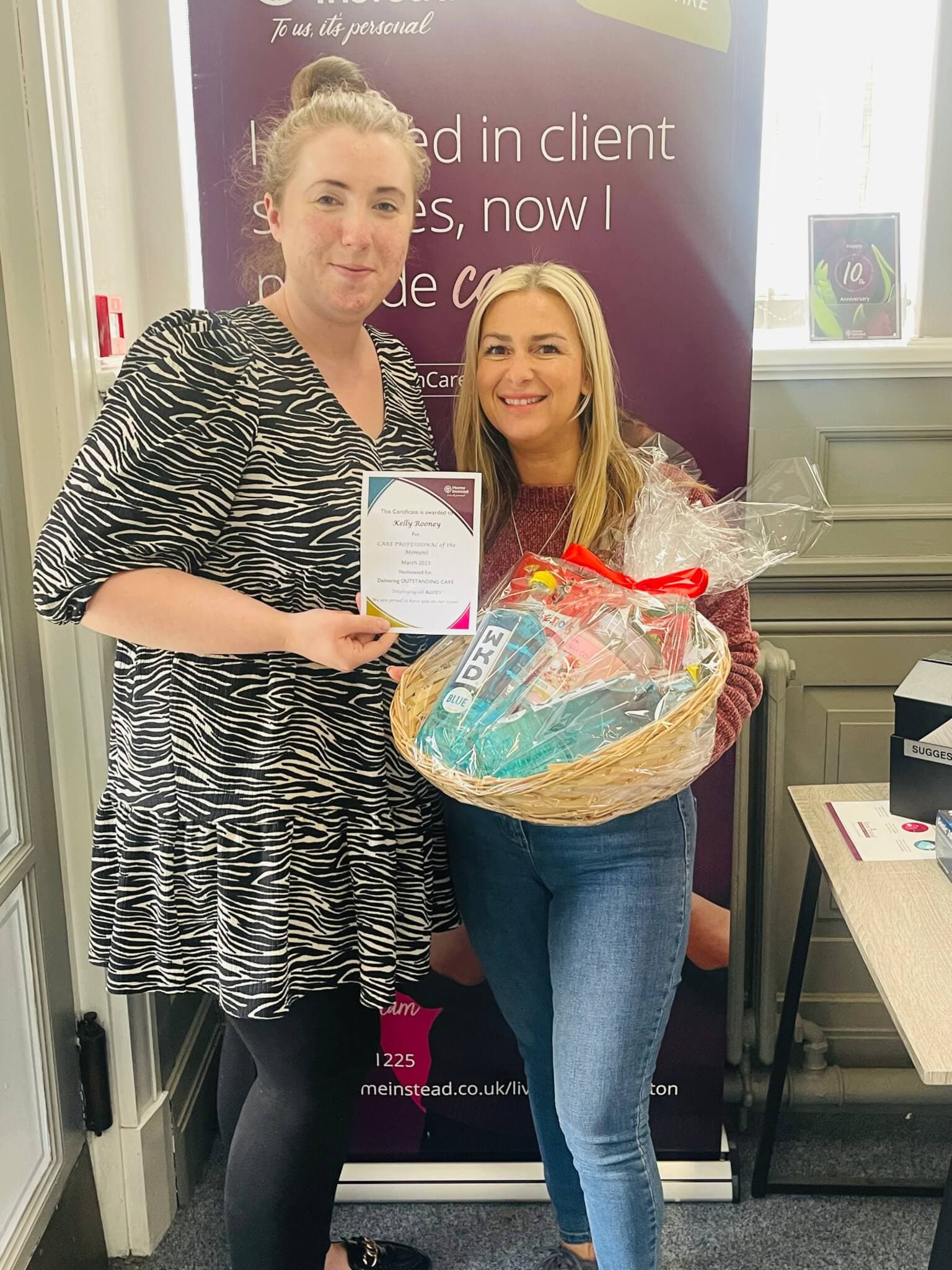 Two women stand together smiling; one holds a certificate, and the other holds a gift basket. - Home Instead