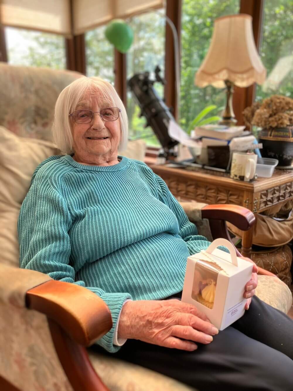 Elderly woman with white hair and glasses, sitting in a cozy chair, holding a small box, smiling contentedly. - Home Instead