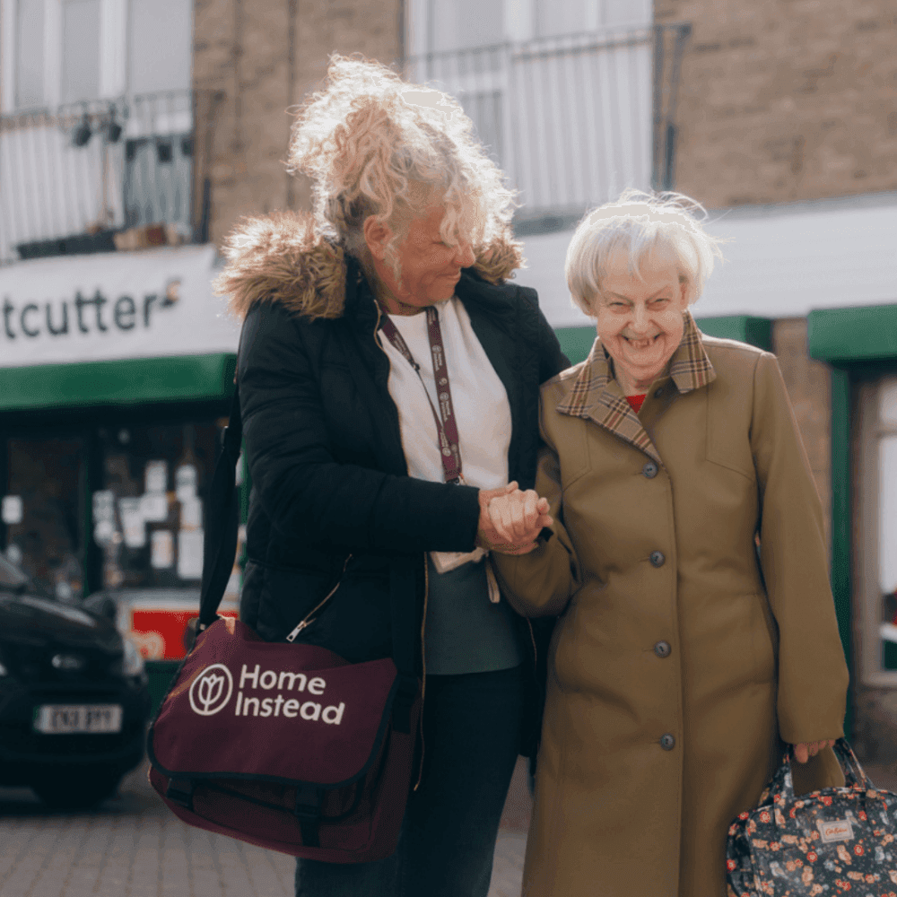 A caregiver assists a smiling elderly woman while holding hands, both standing outdoors in front of shops. - Home Instead