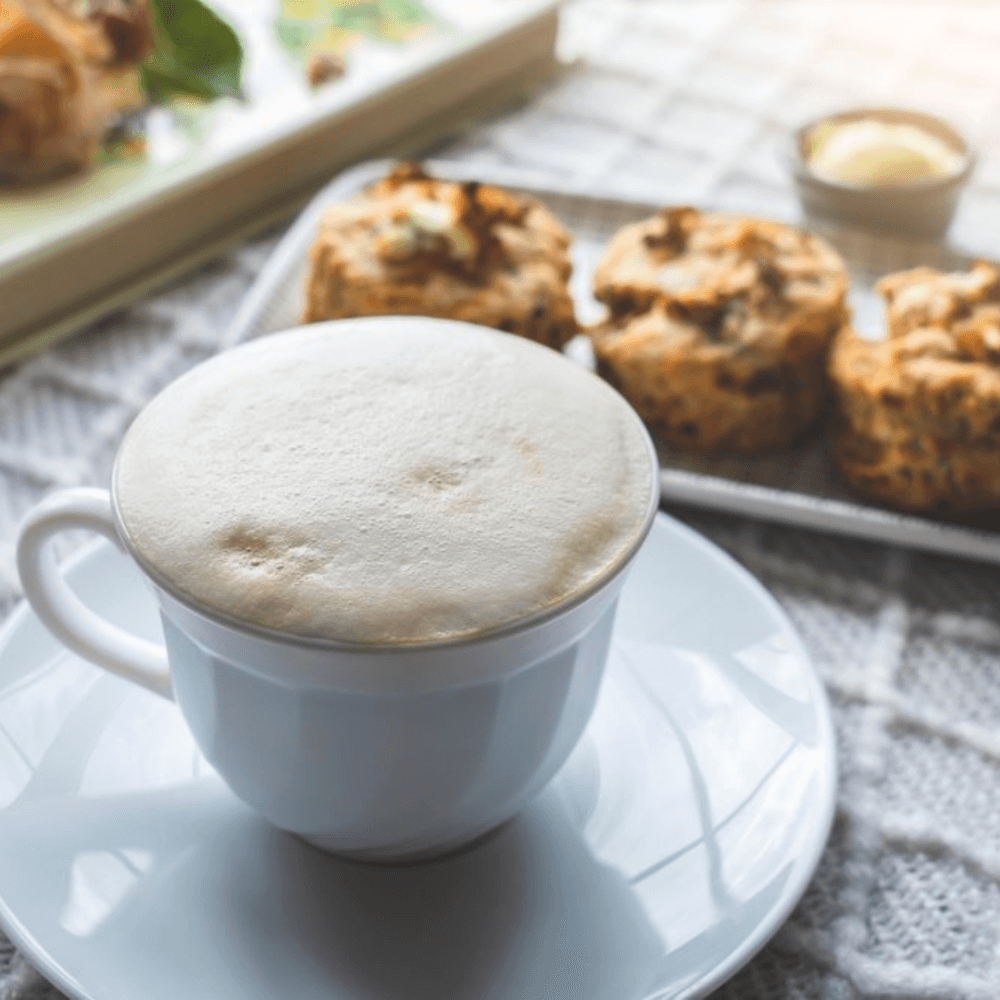 A cappuccino with frothy milk sits on a white saucer, with scones and a small dish of butter in the background. - Home Instead