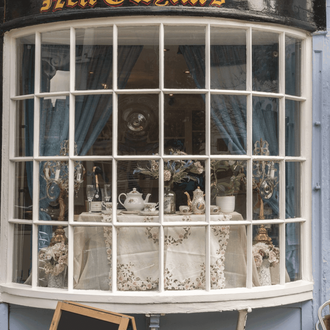 A quaint shop window displays vintage teapots, plants, and a white tablecloth with floral patterns, framed by blue curtains. - Home Instead