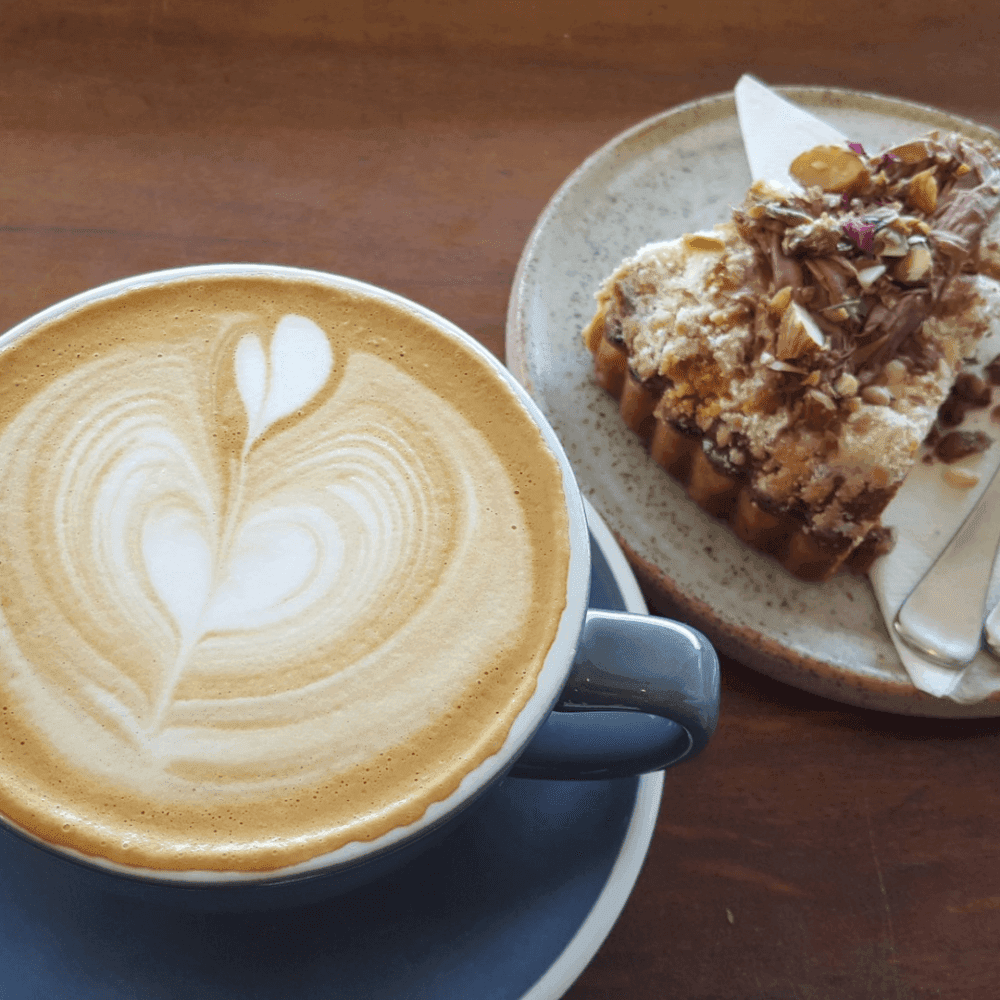 A latte with heart-shaped foam art next to a slice of cake garnished with nuts and icing on a wooden table. - Home Instead