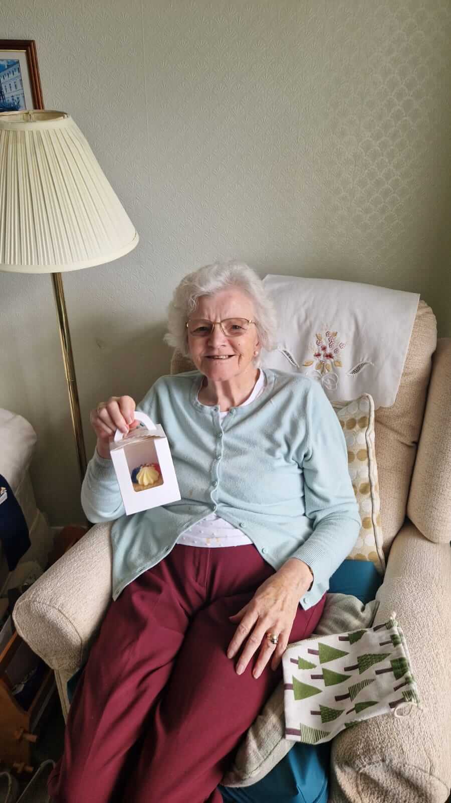 Elderly woman with glasses sitting on a chair, smiling, holding up a small gift box in a cozy, well-lit room. - Home Instead
