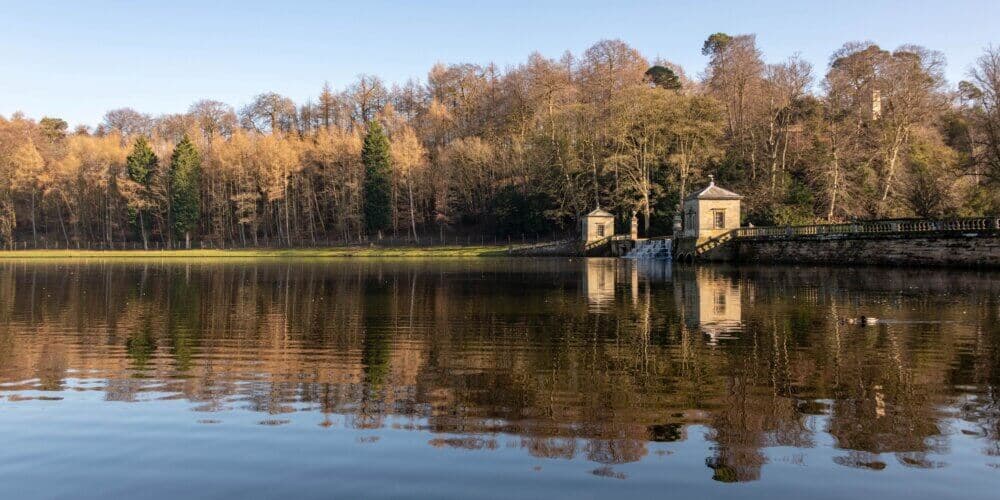 A serene lake reflects a forest and two stone buildings with a small waterfall under a clear blue sky. - Home Instead
